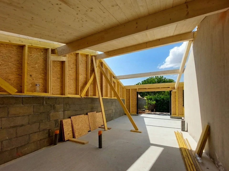 Scène de construction résidentielle avec ouvriers en casques sur chantier maison bois à ossature apparente, échafaudages métalliques, matériaux empilés au sol